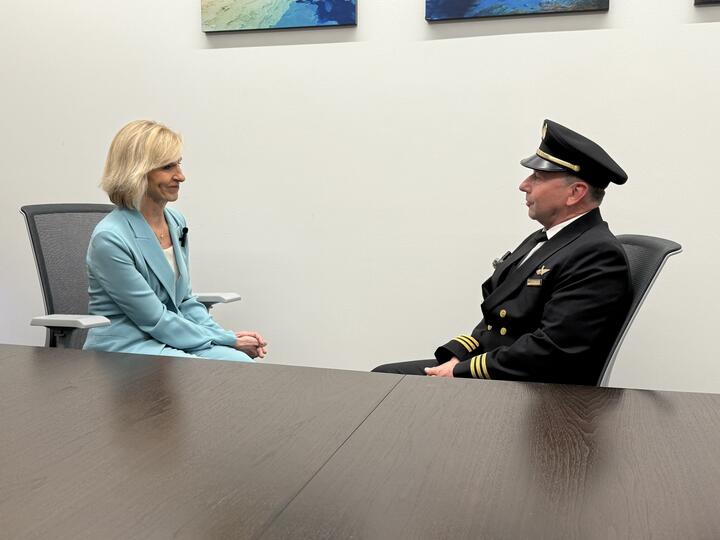 Allison Ausband and Tom Cohan sit facing each other at a conference table, Allison in a business suit and Tom in a pilot uniform.