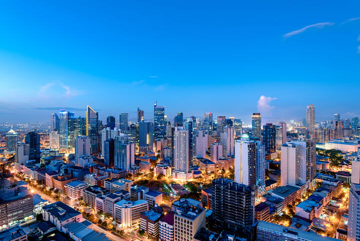 A panoramic view of a modern city skyline at dusk, with illuminated high-rise buildings, busy streets, and a bright blue evening sky.