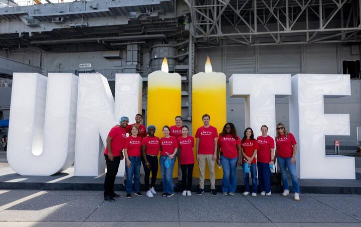 Delta volunteers in red shirts stand in front of a large “UNITE” display with two candle-shaped letters, commemorating 9/11 aboard an aircraft carrier.