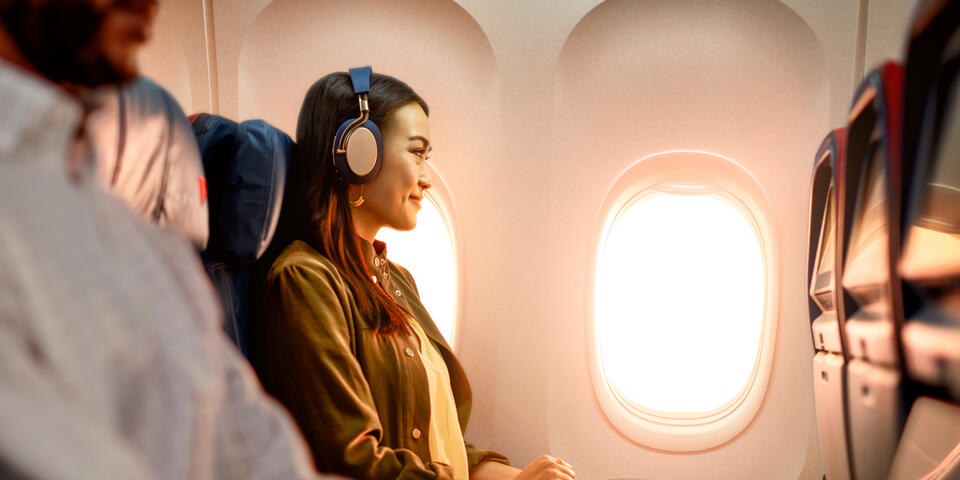 Delta customer on board an airplane wearing headphones sitting by a window, with warm sunlight illuminating the cabin and seatback screens visible.