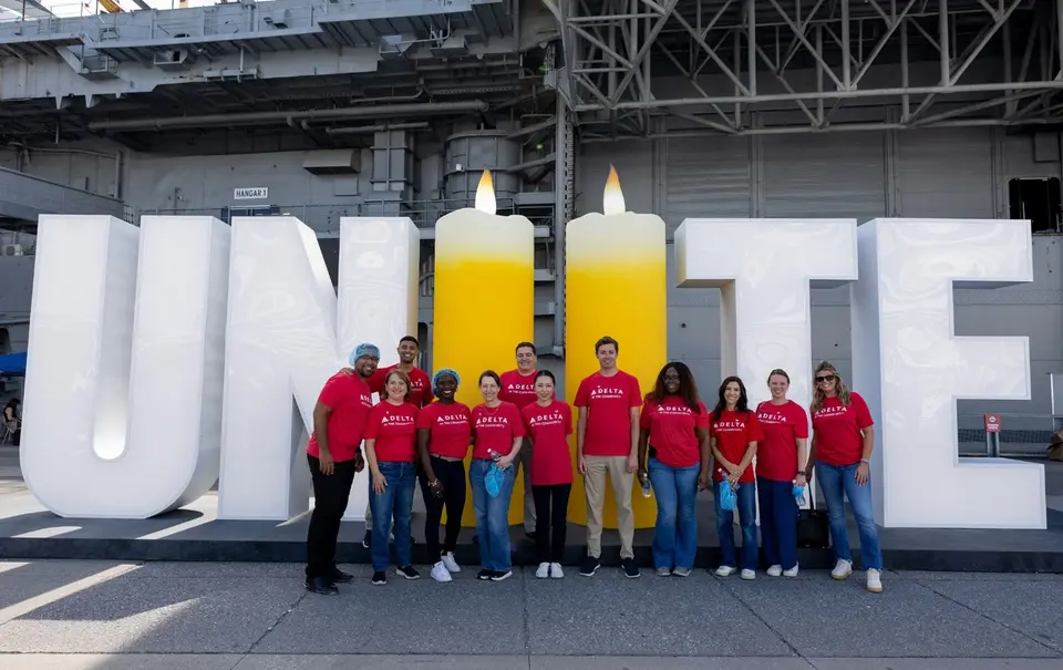 Delta volunteers in red shirts stand in front of a large “UNITE” display with two candle-shaped letters, commemorating 9/11 aboard an aircraft carrier.