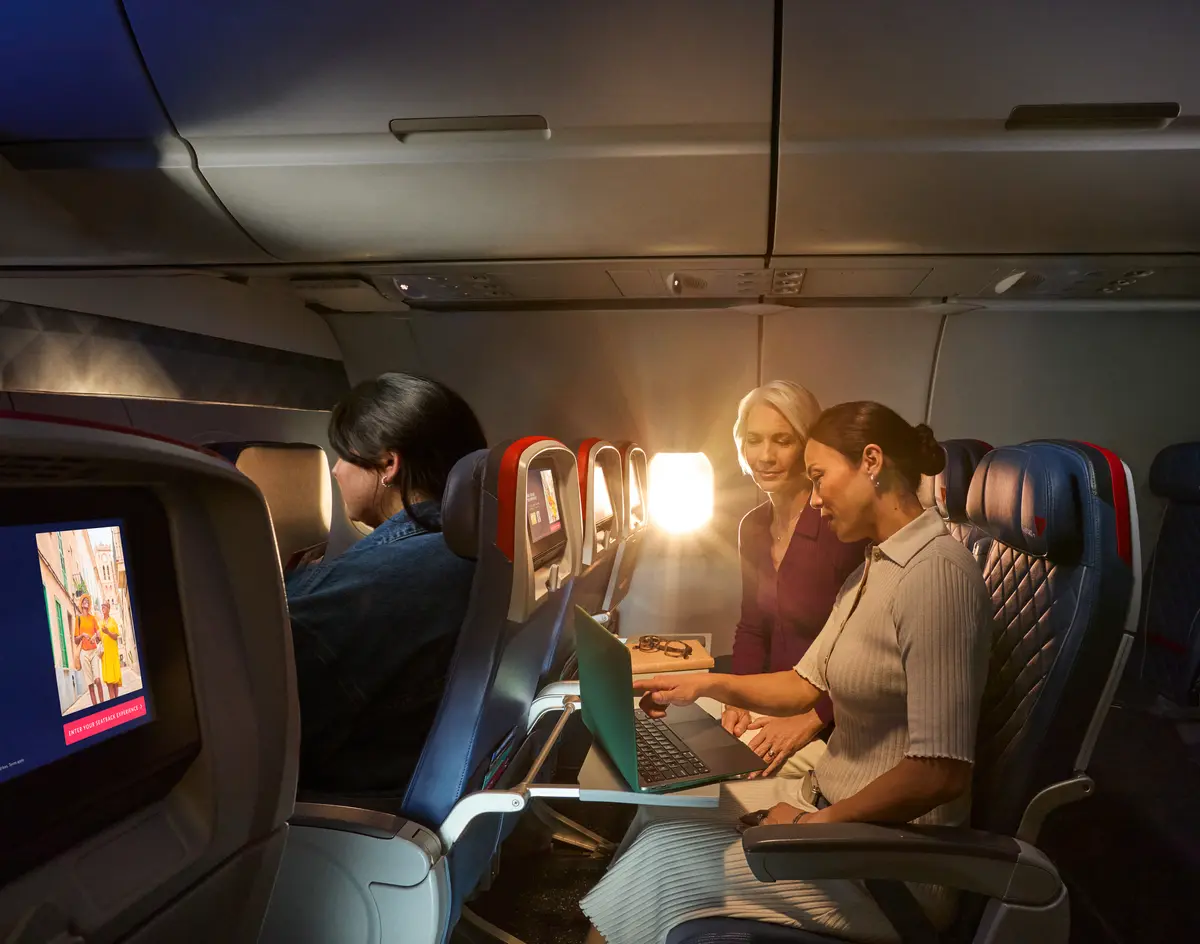 Two female Delta customers on board an aircraft working on laptops, with warm sunlight streaming through the windows.