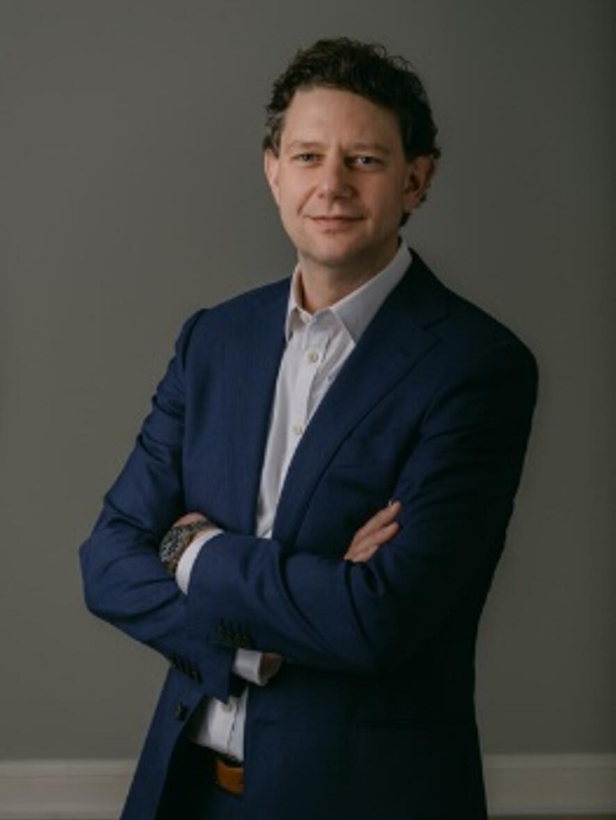 Studio portrait of a person in a dark suit and white shirt, standing with arms crossed against a plain gray background.