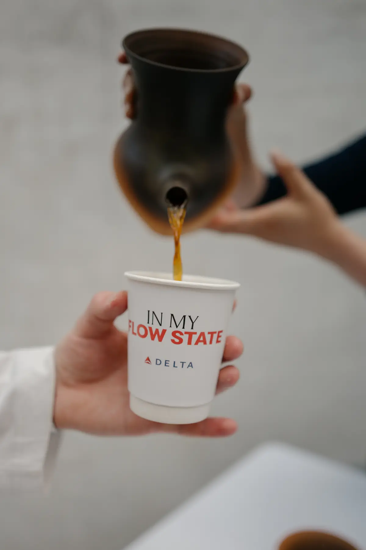 Coffee being poured from a dark ceramic pitcher into a white paper cup printed with “In My Flow State” and the Delta logo.