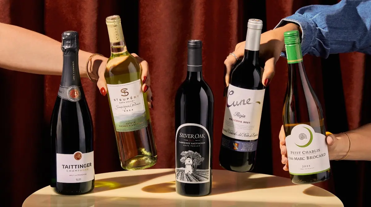 Hands reaching for a selection of sparkling, red and white wine bottles displayed on a table.