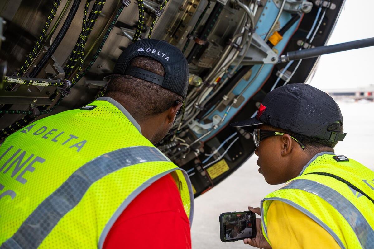 A student and aviation professional examine exposed aircraft engine components while the student records details on a smartphone.