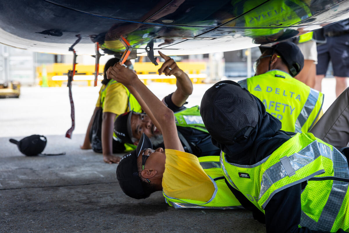 Students in safety vests inspect the underside of a Delta aircraft during a hands-on aviation maintenance learning activity.