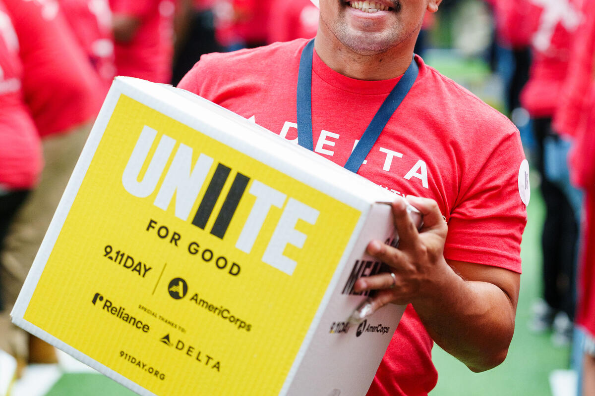 Delta employee wearing a hairnet carries a "Unite for Good" meal kit box at a 9/11 Day volunteer event.