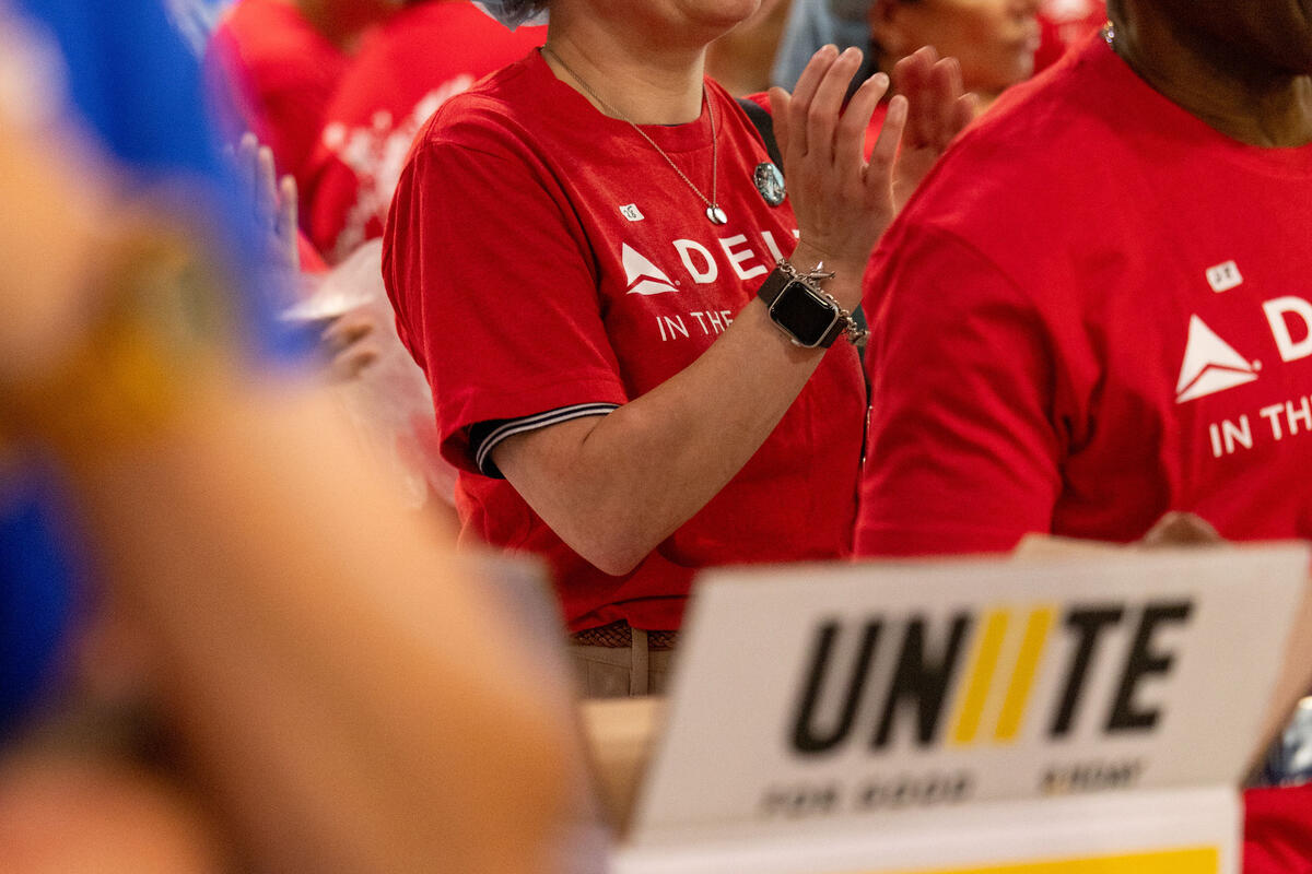 Volunteer in a red Delta in the Community shirt and hairnet applauds during a meal-packing event, with other volunteers and a Unite campaign sign in the background.