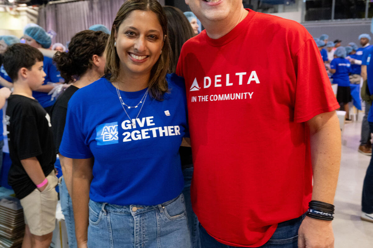 Two volunteers pose together at a large community service event, surrounded by participants preparing meals while wearing hairnets.