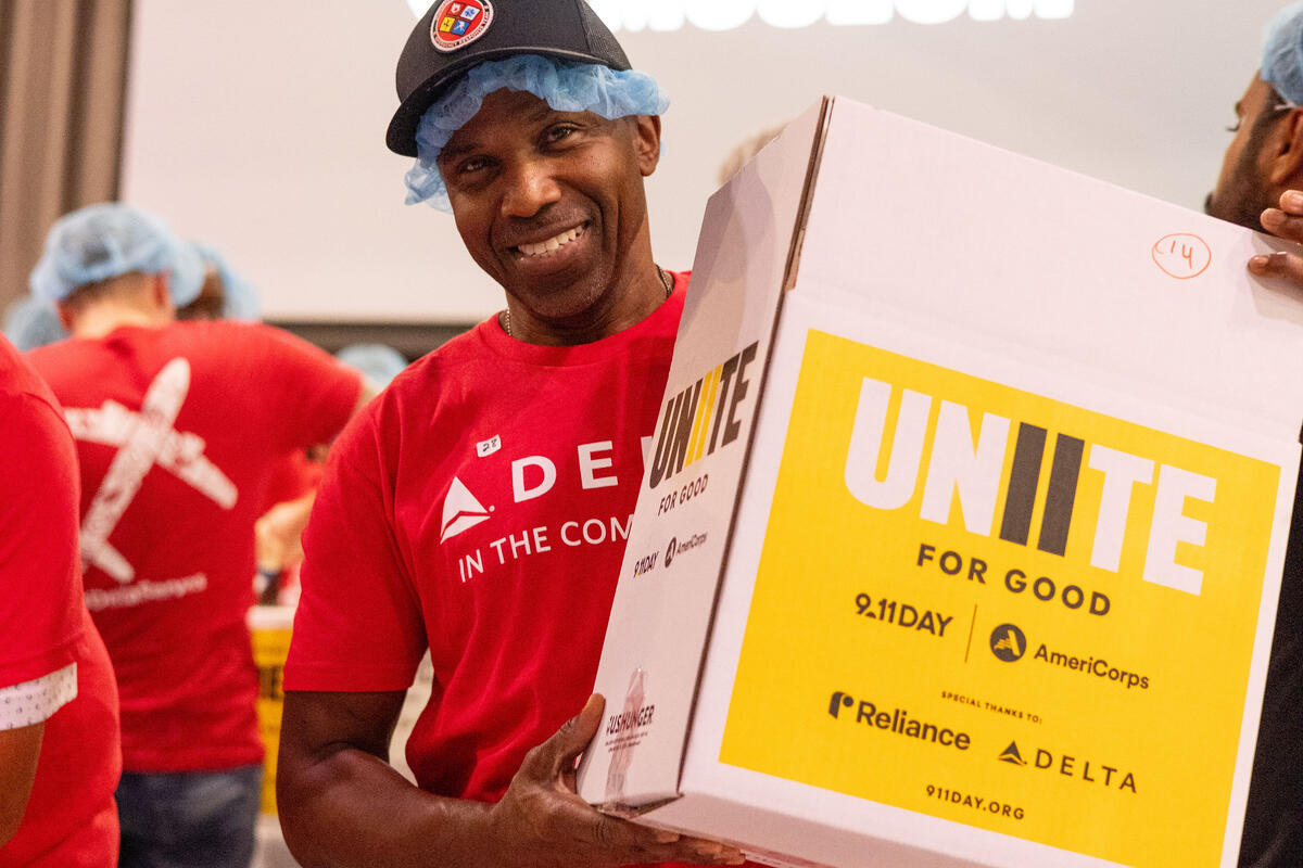 Volunteer in a red Delta in the Community shirt and hairnet carries a Unite for Good meal-packing box during a 9/11 Day service event, with fellow volunteers working in the background.